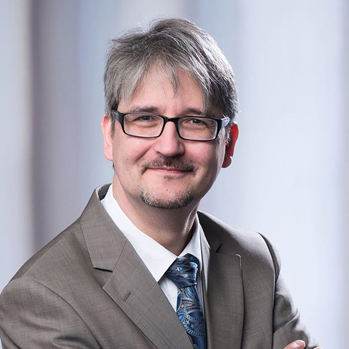 Andreas Sutter, Director von disphere project. Ein Mann mit grauem Haar, einer schwarzen schmalen Brille mit abgerundeten Gläsern, einem Schnauzer sowie Kinnbart. Er trägt einen grauen Anzug mit blauer Krawatte