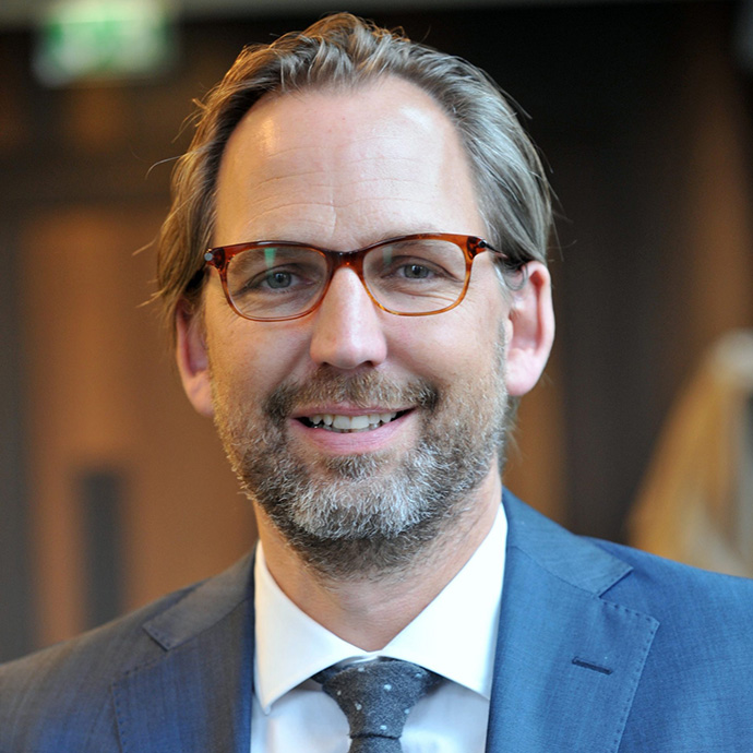 Martin Klein, Rechtsanwalt. Ein Mann mit freundlichem Lächeln, nach hinten gekämmten Haar, einer rot-ovalen Brille, deren obere rechte Ecken spitz zulaufen. Er trägt einen blauen Anzug mit blauer Krawatte.. Der Hintergrund ist verschwommen