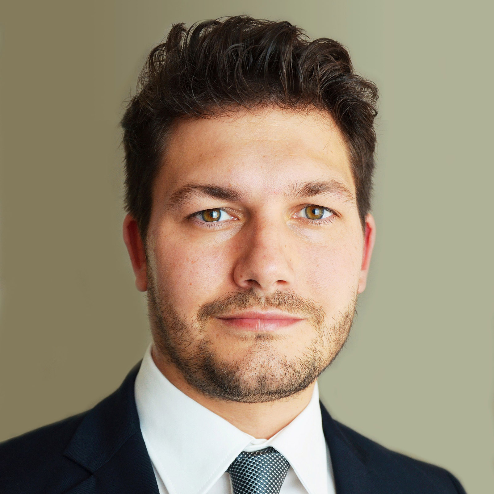 Florian Kölbl, ein junger Mann mit dunklen Haaren und drei Tage Bart. Er trägt einen klasischen Anzug mit Krawatte